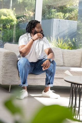 African american man sitting on modern sofa in sunlit living room with garden view