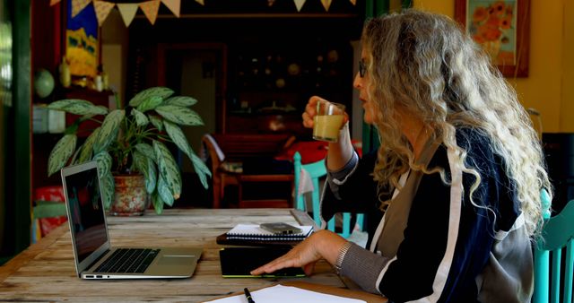 Woman working from home on a laptop, drinking coffee while taking notes. Ideal for illustrating remote work, home office, telecommuting, casual work environment, and productivity at home.