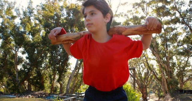 A young boy in a red shirt and dark shorts is exercising with a tree branch on his shoulders, set outdoors in a lush, green forest. Ideal for illustrating healthy habits, kids' activities, outdoor fitness, and nature-based exercises.