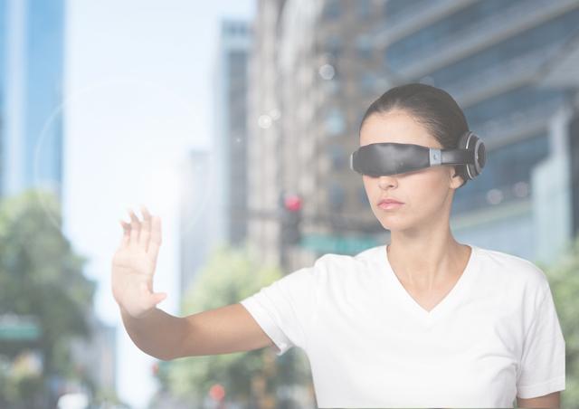 Young woman using a virtual reality headset in an outdoor urban environment. She is engaged in an interactive experience, pictured lifting her hand in a gesture as if interacting with a virtual interface. Bright background with city buildings and trees giving a futuristic feel. Ideal for use in content related to technology, innovation, augmented reality, and modern lifestyle.