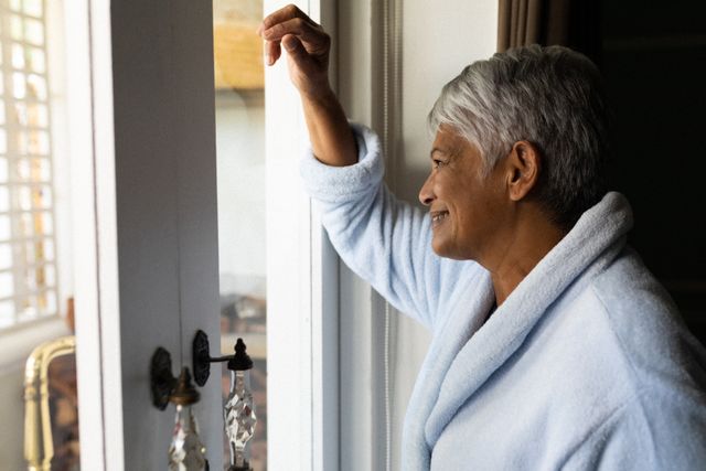 Senior woman standing by a window in a bedroom, dressed in a robe, looking outside thoughtfully. Ideal for use in articles or advertisements about retirement, self-isolation, wellness, and the impact of the coronavirus pandemic on the elderly. Can also be used in content related to peaceful mornings and home life.