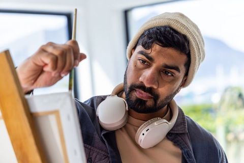 Indian artist painting on canvas in sunlit studio wearing beanie and headphones