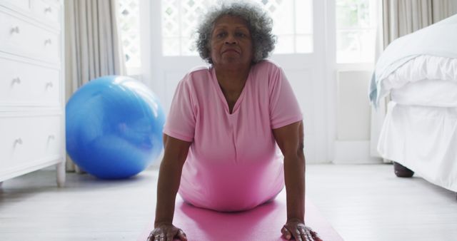 Senior woman practicing yoga on pink mat at home, with blue exercise ball in background. Ideal for promoting home fitness routines, senior health and wellness, active aging, balanced lifestyle, and exercises for maintaining flexibility and strength among the elderly community.