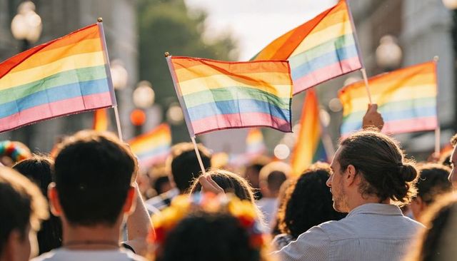 Diverse crowd waving rainbow pride flags during sunlit city parade celebrating pride