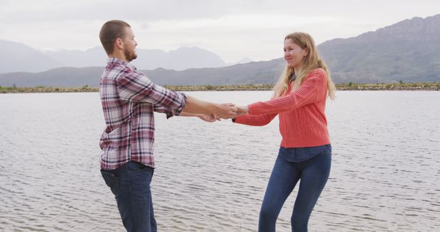 A young couple enjoys a fun moment dancing by a picturesque lake with mountains in the background. This image captures their joyful expressions, highlighting their close bond and love for the outdoors. Ideal for use in relationship blogs, travel websites, outdoor adventure promotions, and lifestyle magazines.