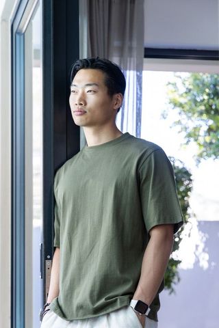 Young asian man standing by sunlit sliding door wearing olive t-shirt and smartwatch