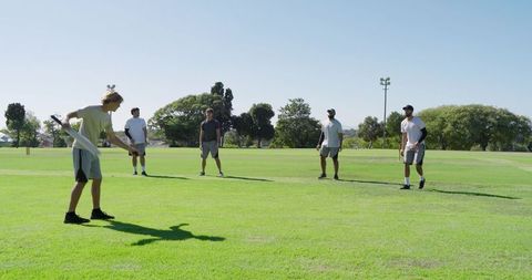 Diverse male teammates playing cricket on sunny park field batting and fielding