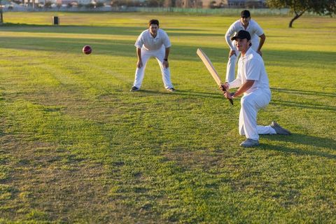 Batsman kneeling for sweep shot with teammates fielding on cricket outfield at sunset