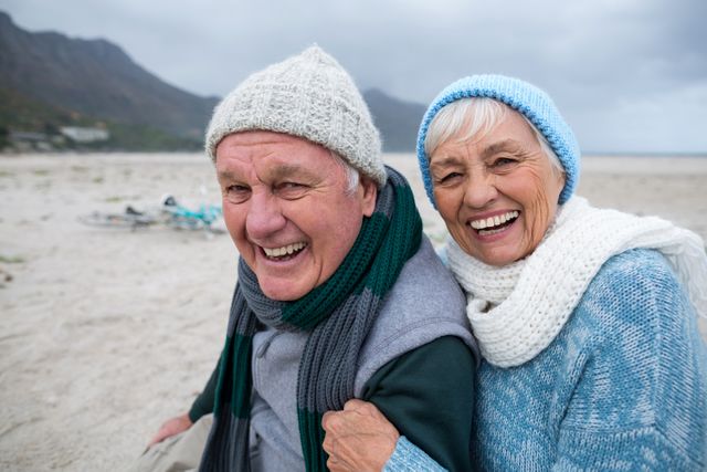 Senior couple enjoying a day at the beach during winter, dressed warmly in scarves and beanies. Ideal for use in advertisements, retirement planning brochures, health and wellness campaigns, and lifestyle blogs focusing on senior living and outdoor activities.