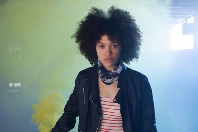 Transgender woman with afro hair holding a blue smoke grenade in an empty parking garage. She is wearing a leather jacket and a striped shirt, symbolizing gender expression and diversity. This image can be used for campaigns promoting gender identity, empowerment, and pride.
