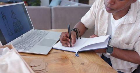 Fashion designer taking notes at wooden desk with laptop displaying garment sketches
