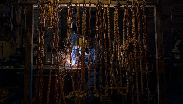 Welding craftsman sparking metal amid hanging chains and heavy ropes in dim workshop
