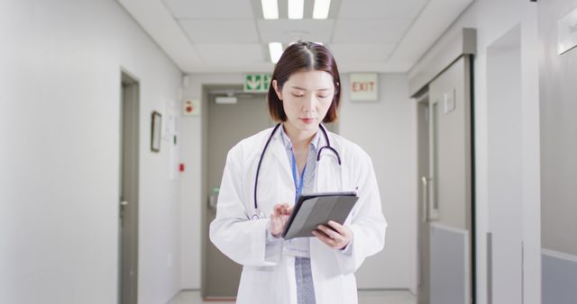 Image of asian female doctor walking in hospital corridor, looking at tablet, copy space. Hospital, medical and healthcare services.