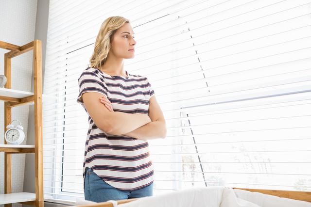 This image depicts a woman standing indoors, looking out a window with a thoughtful expression. She is wearing a casual striped shirt and has blonde hair. The natural light coming through the blinds adds a serene and peaceful atmosphere. This image can be used for themes related to contemplation, introspection, mental health, solitude, and peaceful moments at home.