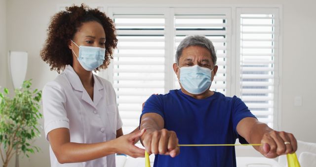 Healthcare worker assists elderly man with physical therapy using a resistance band. Both are wearing face masks for health and safety. Ideal for use in articles about elderly care, physical rehabilitation, pandemic patient care, or home healthcare services.