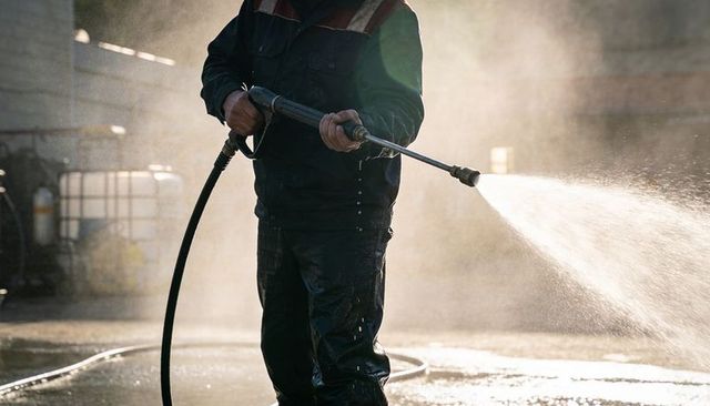 Industrial worker power-washing concrete yard with high-pressure wand and protective suit