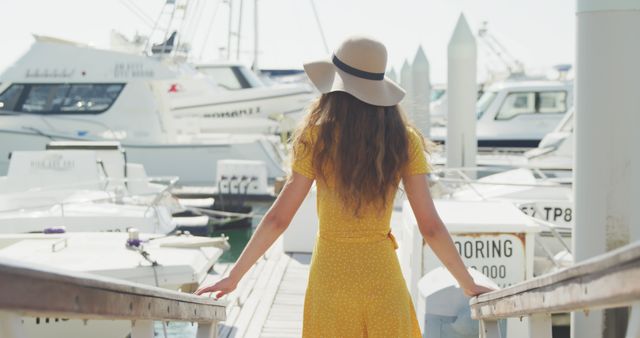 Rear view of caucasian teenage girl in hat and yellow dress standing on jetty by boats on sunny day. Leisure, free time, travel and vacations.
