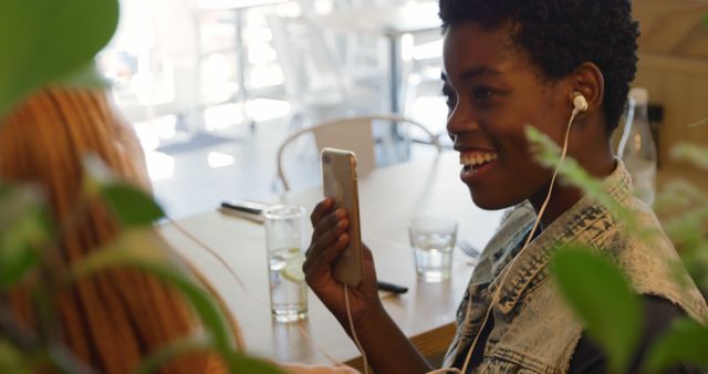 This image features a young African American woman smiling while listening to music on her smartphone and chatting with a friend at a cafe. It can be used in promotional materials for modern lifestyle blogs, social media campaigns, friendship and leisure activities, or cafes advertising a cozy ambience for catching up.