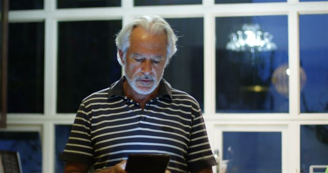 Senior man with white hair and beard reading on a digital tablet at home during the evening. Relaxing at home, using modern technology for reading or browsing the internet. Suitable for articles and advertisements related to senior lifestyle, technology use among older adults, or home and lifestyle features.