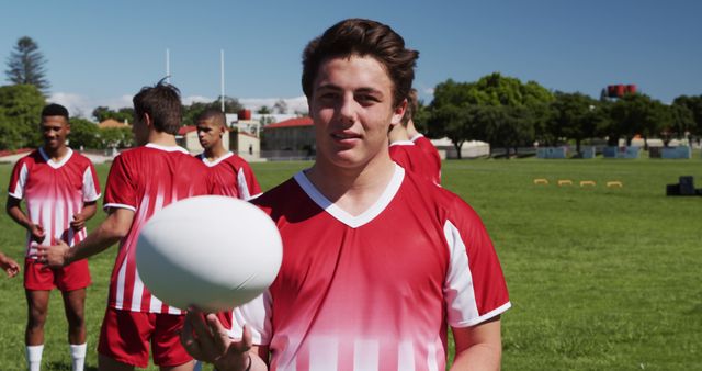 Teenage boy in red and white jersey holding a rugby ball with teammates in the background. Ideal for use in advertisements or articles about youth sports, teamwork, fitness, and outdoor activities.