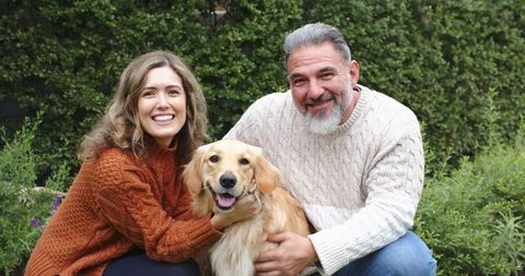 Smiling couple kneeling with golden retriever in cozy garden wearing knit sweaters