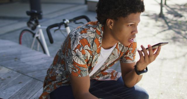 Young man sitting outdoors using voice commands on smartphone, dressed in casual clothing with a bicycle in the background. Ideal for content related to technology, smart devices, young adult lifestyle, and modern communication.