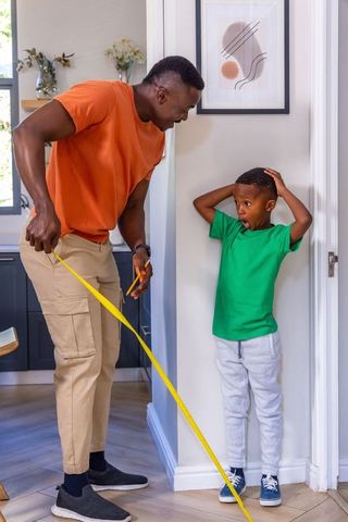 African american father measuring wall with tape measure while son reacts in surprise