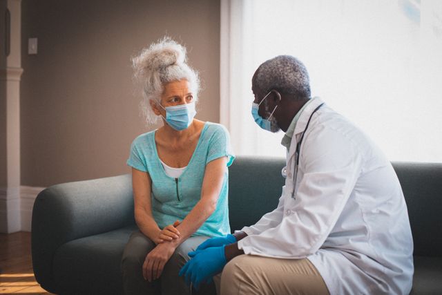 African american senior male doctor talking to female patient at home both wearing face masks. staying at home in isolation during quarantine lockdown.