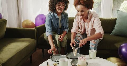 Two African American women sharing cake on green sofa during cozy at-home celebration