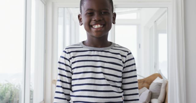 Young African American boy wearing striped shirt, smiling. Bright, modern living room with natural light. Perfect for articles on child happiness, family life, and advertising children's clothing.