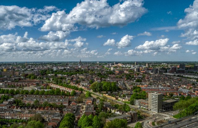 Wide aerial view of a city skyline showcasing residential buildings, greenery, and urban infrastructure under a bright blue sky with scattered clouds. This image is ideal for use in real estate promotions, urban development presentations, and travel brochures highlighting city architecture and landscapes.