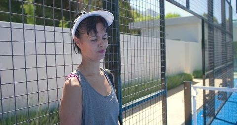 Mature woman standing by fenced blue court wearing white visor and athletic grey tank