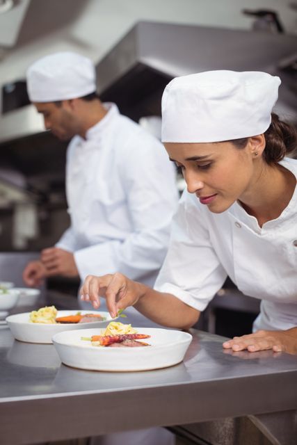 Female chef meticulously garnishing a dish in a professional restaurant kitchen, highlighting precision and attention to detail. Another chef working in the background, showing teamwork and collaborative cooking environment. Perfect for content related to culinary arts, professional cooking, restaurant operations, and hospitality training materials.