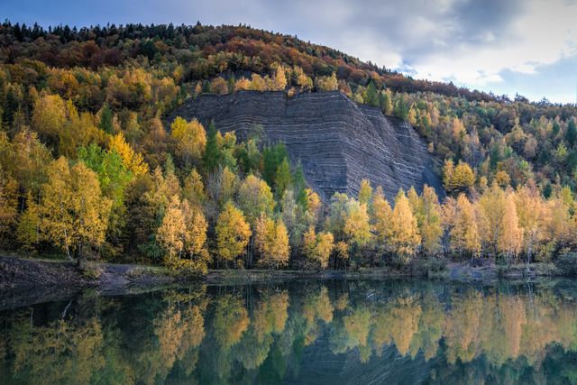 Colorful autumn foliage of a forest by a tranquil lake, with tree reflections creating mirror effect on water. Striated rocky cliff adds to picturesque scenery under a clouded sky. Ideal for promoting outdoor activities, travel destinations, nature tours, landscape photography, and seasonal calendars.