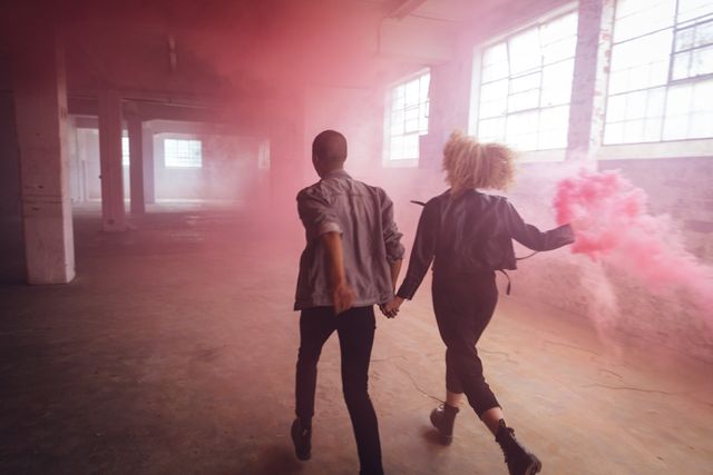Rear view of a hip young biracial man and a hip young Caucasian woman in an empty warehouse, holding hands and running, the woman holding a hand grenade, with pink smoke.