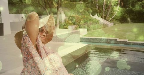 Senior woman relaxing by mosaic pool adjusting sunhat and sunglasses in backyard oasis