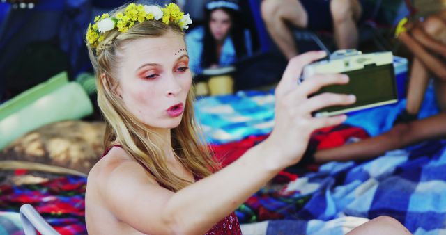 A young Caucasian girl is taking a selfie with a vintage camera at an outdoor gathering, with copy space. Her floral headband and face paint suggest a festive or bohemian event.