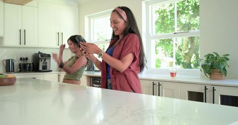 Multiethnic friends dancing and texting in sunlit modern kitchen with marble island