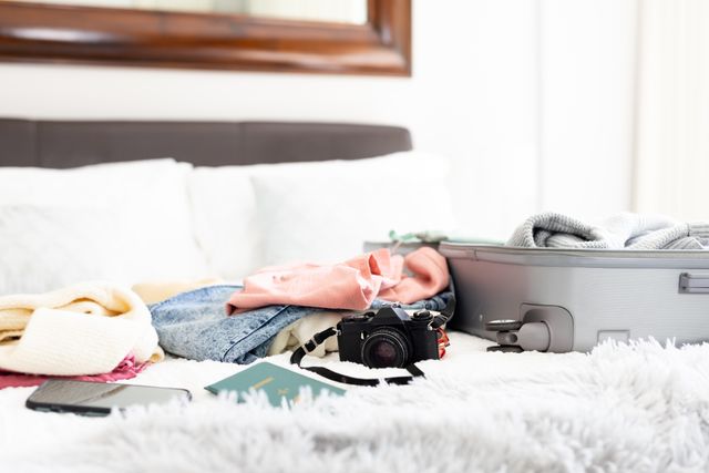 Image of camera, smartphone, passport, folded clothes and open suitcase on bed at home, copy space. Travel, holiday, preparation and lifestyle concept.