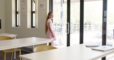 Sunlit classroom student standing by glass doors wearing pink gingham dress minimalist interior