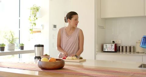 Sunlit woman slicing bananas and strawberries on modern kitchen island, healthy breakfast