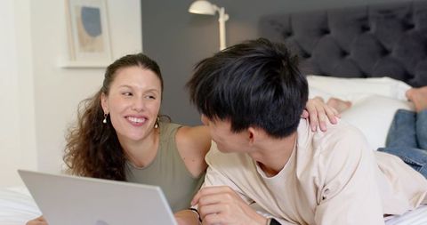 Mixed-race couple relaxing on bed with laptop sharing intimate cozy moment