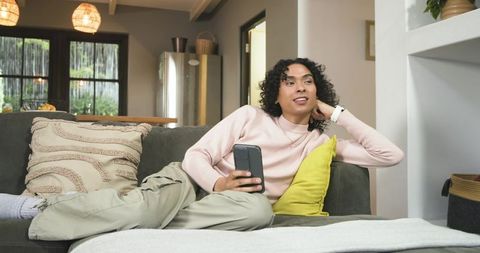 Nonbinary asian person relaxing on sofa holding smartphone in cozy contemporary room