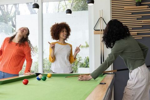 Young women playing pool in sunlit recreation room laughing and socializing