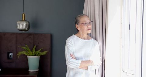 Contemplative Senior Asian Woman by Window in Sunlit Bedroom