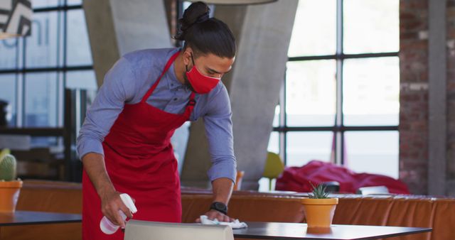 Restaurant worker in red apron and mask is cleaning table with spray bottle inside modern restaurant. Useful for topics related to health, safety, hygiene, COVID-19 precautions, customer service, and daily operations in hospitality industry.