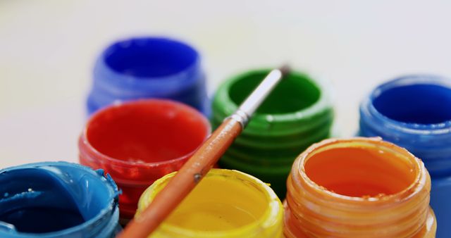 Open jars of vibrant watercolor paints are arranged in a row, with a paintbrush resting atop, with copy space. This setup is typical for an artist's workspace, ready for a creative painting session.