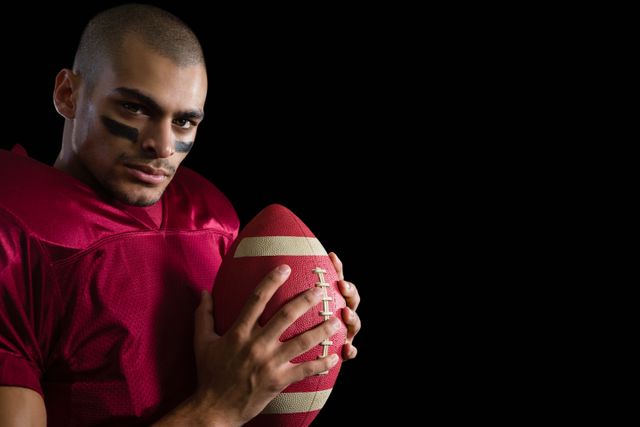 Determined American football player holding a football with both his hands against a black background