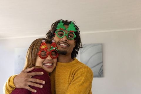 Couple hugging in cozy christmas sweaters wearing festive novelty glasses, sharing laughter