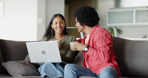 Multiracial Couple Sitting on Sofa with Laptop and Coffee Sharing Cozy Conversation